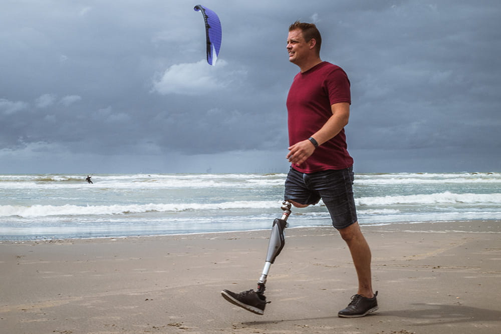 A man with an endo-exo prosthesis is walking along the beach and behind him is a kitesurfer in the water.