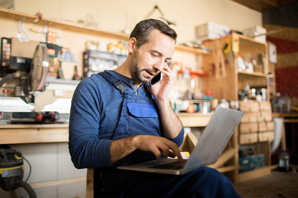 A craftsman sits in front of his laptop with a phone to his ear.
