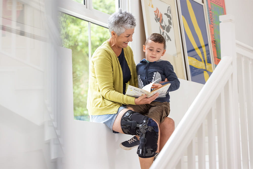 An elderly lady sits on a staircase ledge and reads a book to her grandson. She is wearing a leg orthosis.