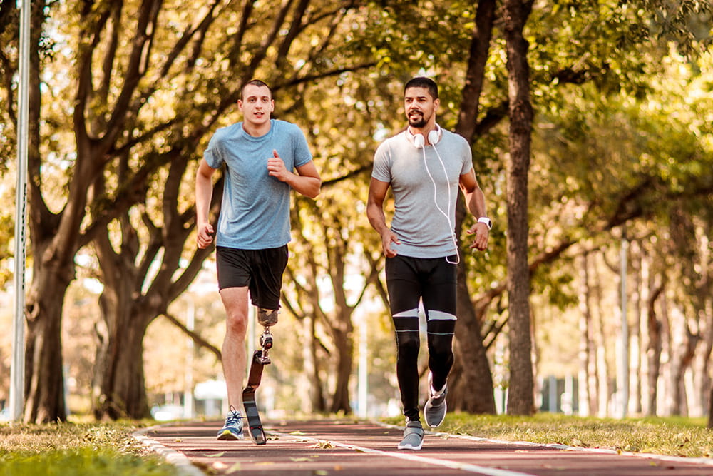 Two joggers are running on a sports track in autumn. One of the runners wears a lower leg prosthesis.