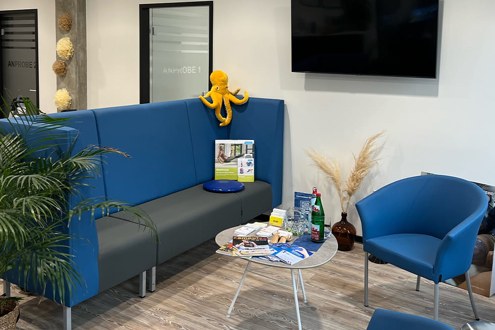 A view into the waiting room of the TOH Technical Orthopaedics Hamburg. There is a blue couch and 2 blue armchairs, a small table and a picture of Hamburg on the wall.