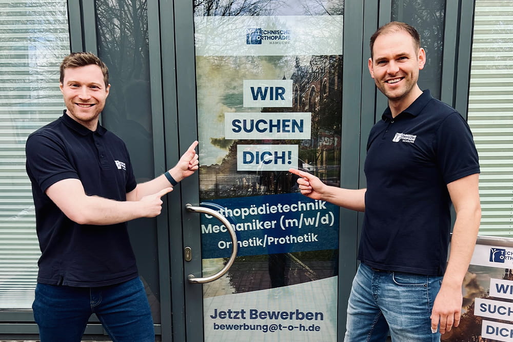 Die beiden Inhaber Maximilian und Florian stehen vor einer gläsernen Ladentür mit einem Poster, auf dem für neue Mitarbeiter gesucht wird.