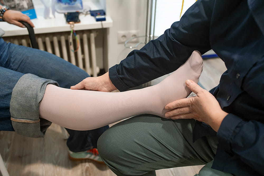 Eine Kundin hat ihr Bein auf dem der Sanitätshausmitarbeiterin und ihr wird ein Kompressionsstrumpf angezogen.