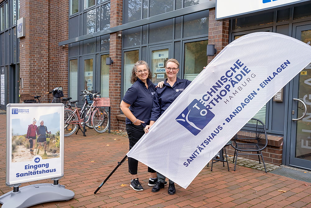 Zwei Mitarbeiter der TOH Technischen Orthopädie Hamburg stehen vor dem Laden und halten eine Fahne in der Hand.