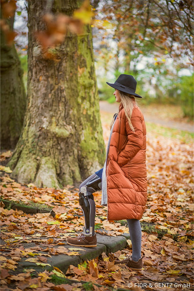 Eine Frau steht im Herbst im Waldmit einer auffälligen orangen jacke und einem dunklen Hut und zeigt dabei ihre Ganzbein-Orthese von Fior und Gentz.