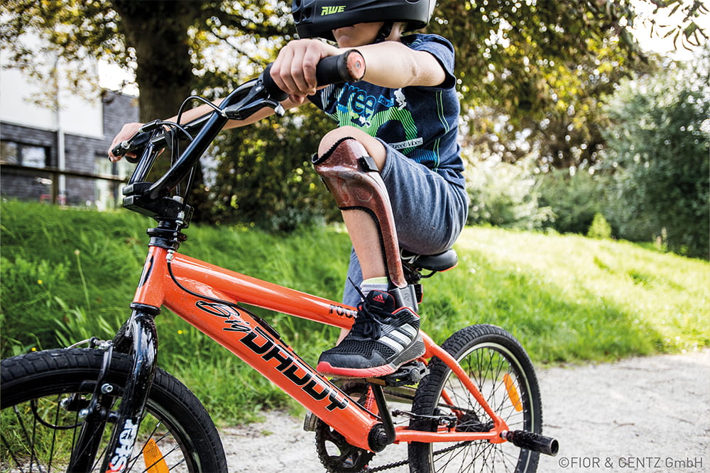 Ein Junge auf einem orangen Fahrrad trägt eine Beinorthese von Fior und Gentz.